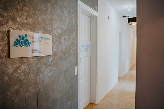 Flur mit dem Praxisschild der Tropenordination an einer grauen Wand neben einer weißen Eingangstür.